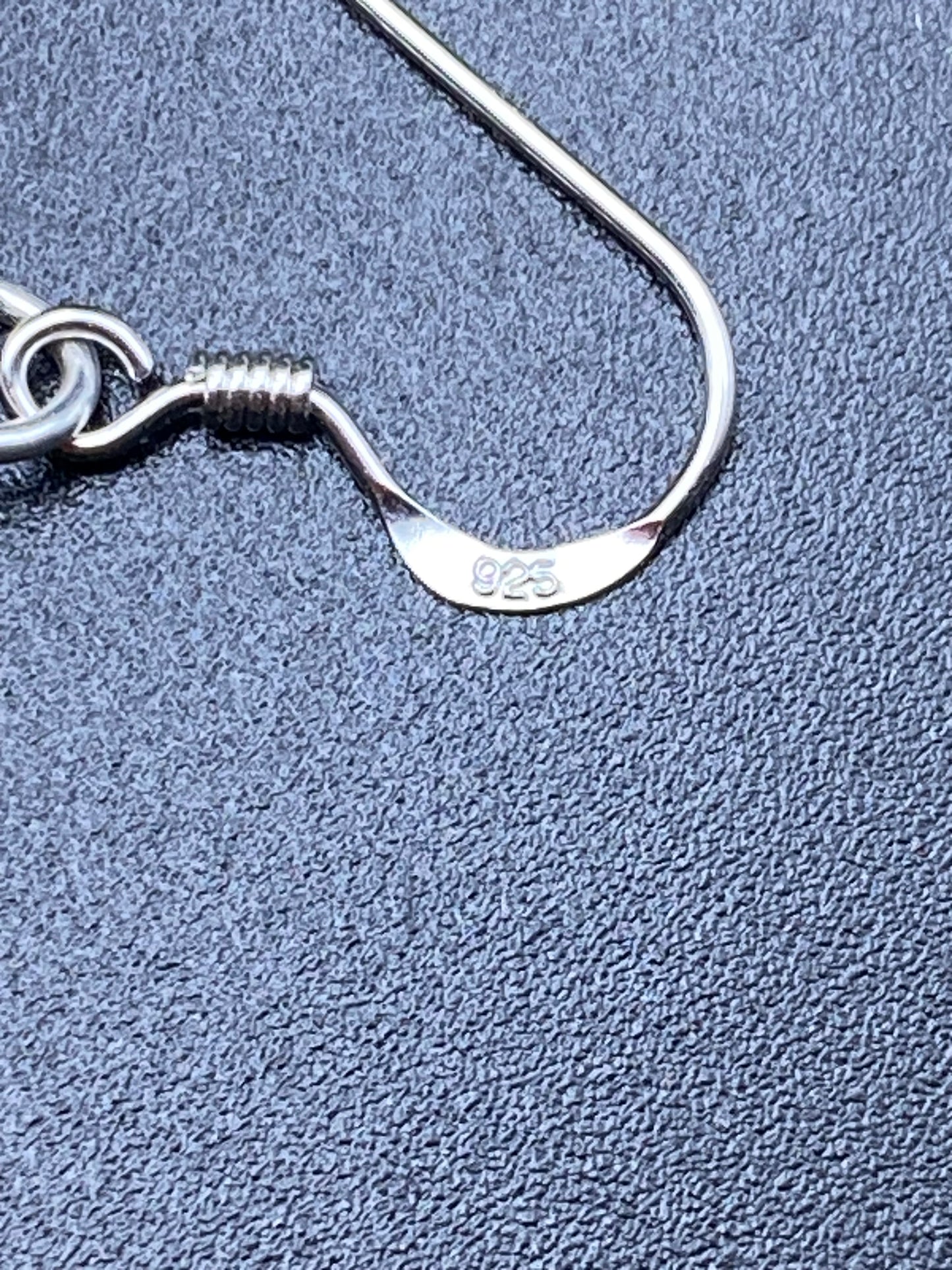 May include: Close-up of sterling silver earring hooks. The hooks are made of shiny metal and have a simple, elegant design. The metal is marked with '925', indicating that it is sterling silver. The earring hooks are shown against a textured, dark gray background.