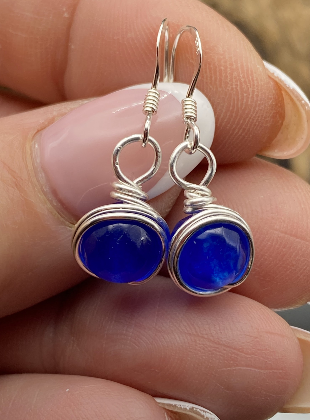 Close-up of dainty sapphire blue single bead drop earrings wrapped in silver, displayed on a soft neutral background.