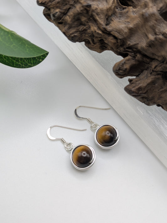 Silver earrings featuring round Tiger’s Eye gemstones with golden-brown chatoyancy, displayed on a crystal slab.
