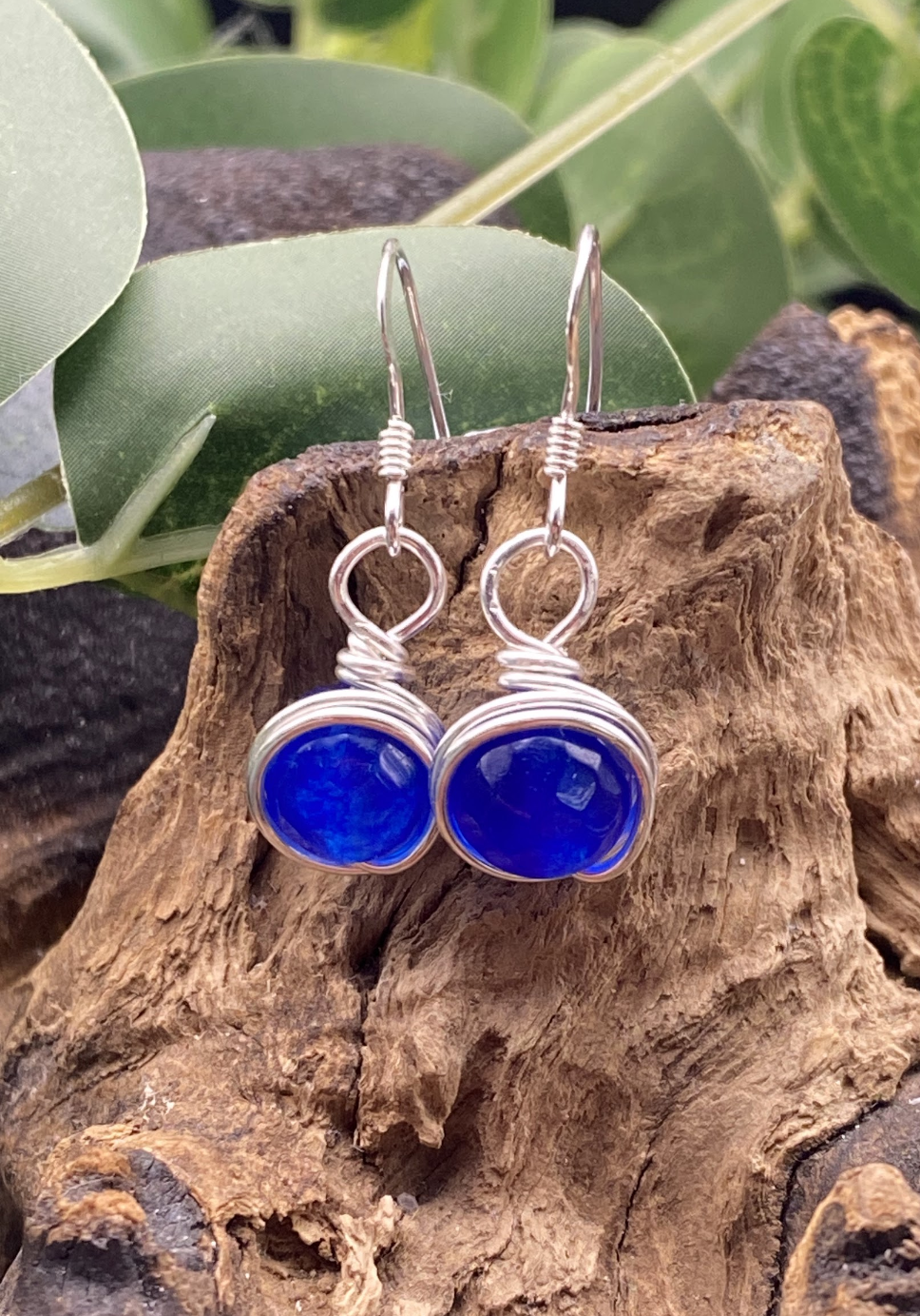 Close-up of dainty sapphire blue single bead drop earrings wrapped in silver, displayed on a soft neutral background.