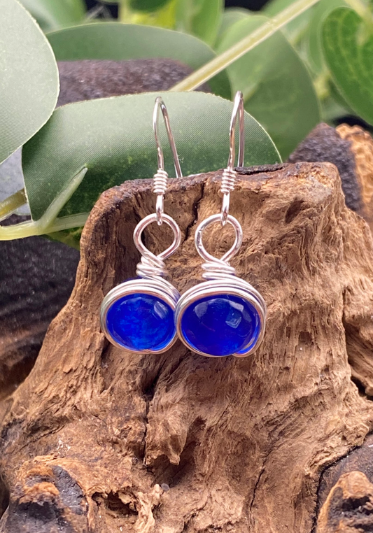 Close-up of dainty sapphire blue single bead drop earrings wrapped in silver, displayed on a soft neutral background.