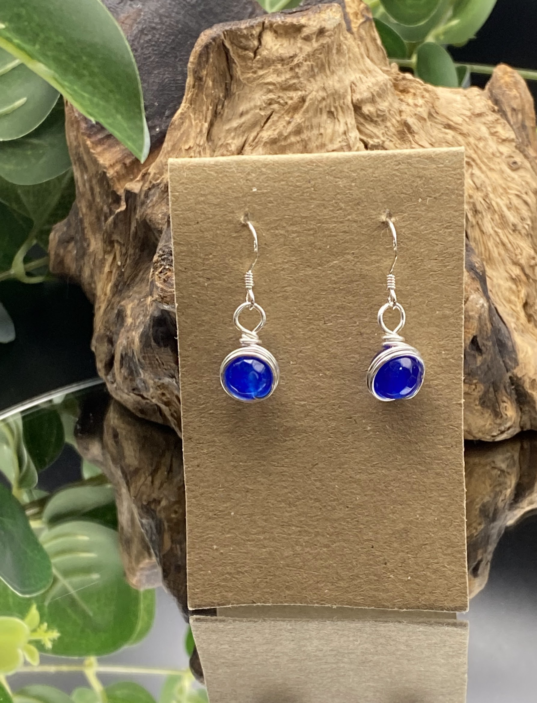 Close-up of dainty sapphire blue single bead drop earrings wrapped in silver, displayed on a soft neutral background.