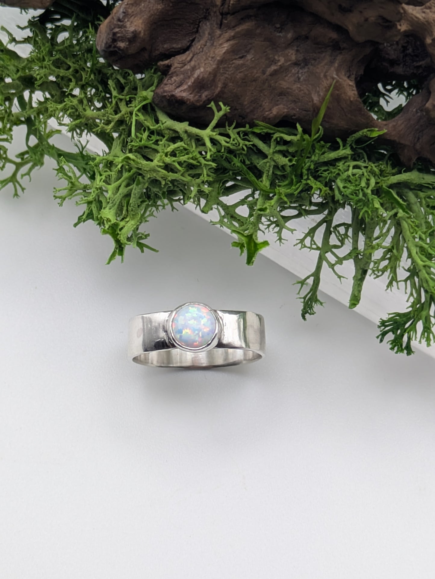 Silver ring with a central opal stone on a white surface with green foliage in the background