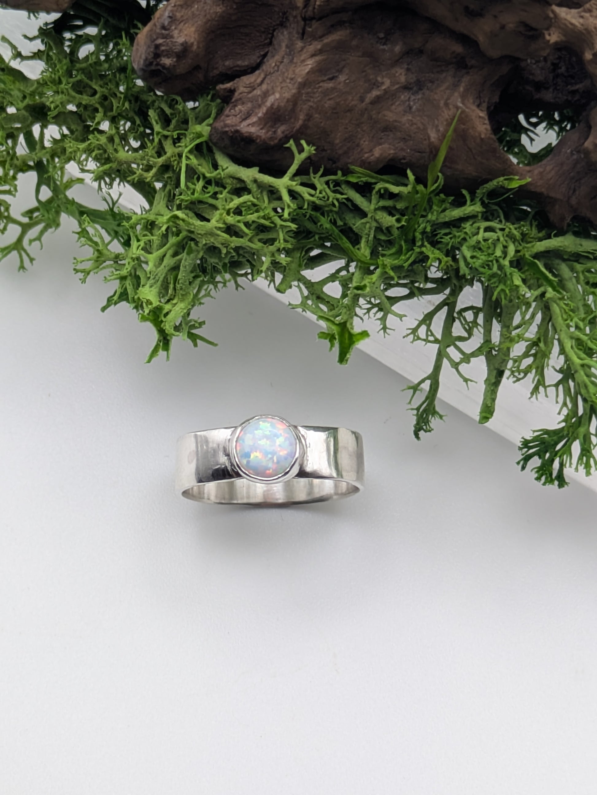 Silver ring with a central opal stone on a white surface with green foliage in the background