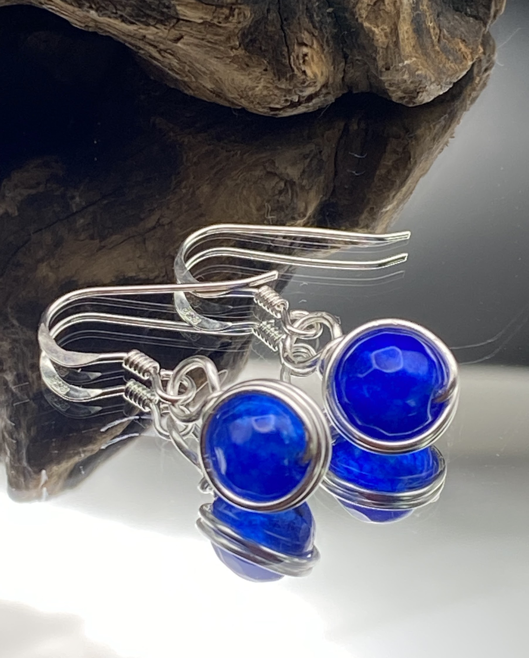 Close-up of dainty sapphire blue single bead drop earrings wrapped in silver, displayed on a soft neutral background.