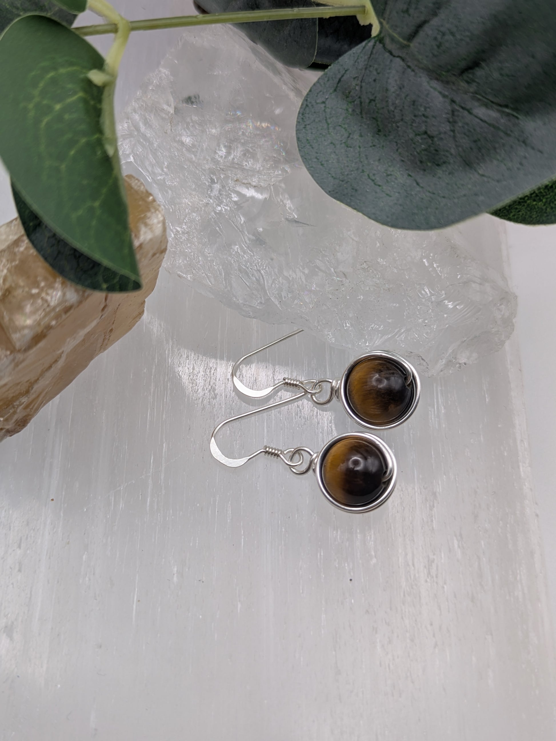 Silver earrings featuring round Tiger’s Eye gemstones with golden-brown chatoyancy, displayed on a crystal slab.