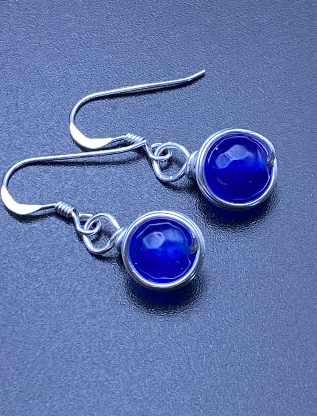 Close-up of dainty sapphire blue single bead drop earrings wrapped in silver, displayed on a soft neutral background.