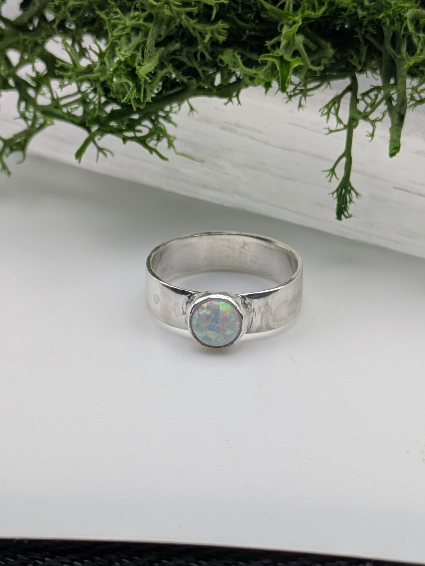 Silver ring with an opal stone on a white surface with greenery in the background