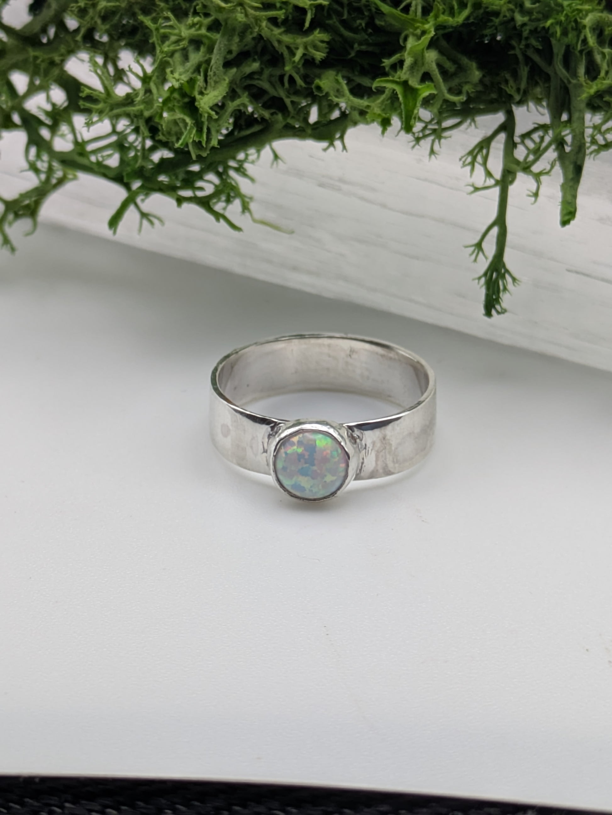 Silver ring with an opal stone on a white surface with greenery in the background