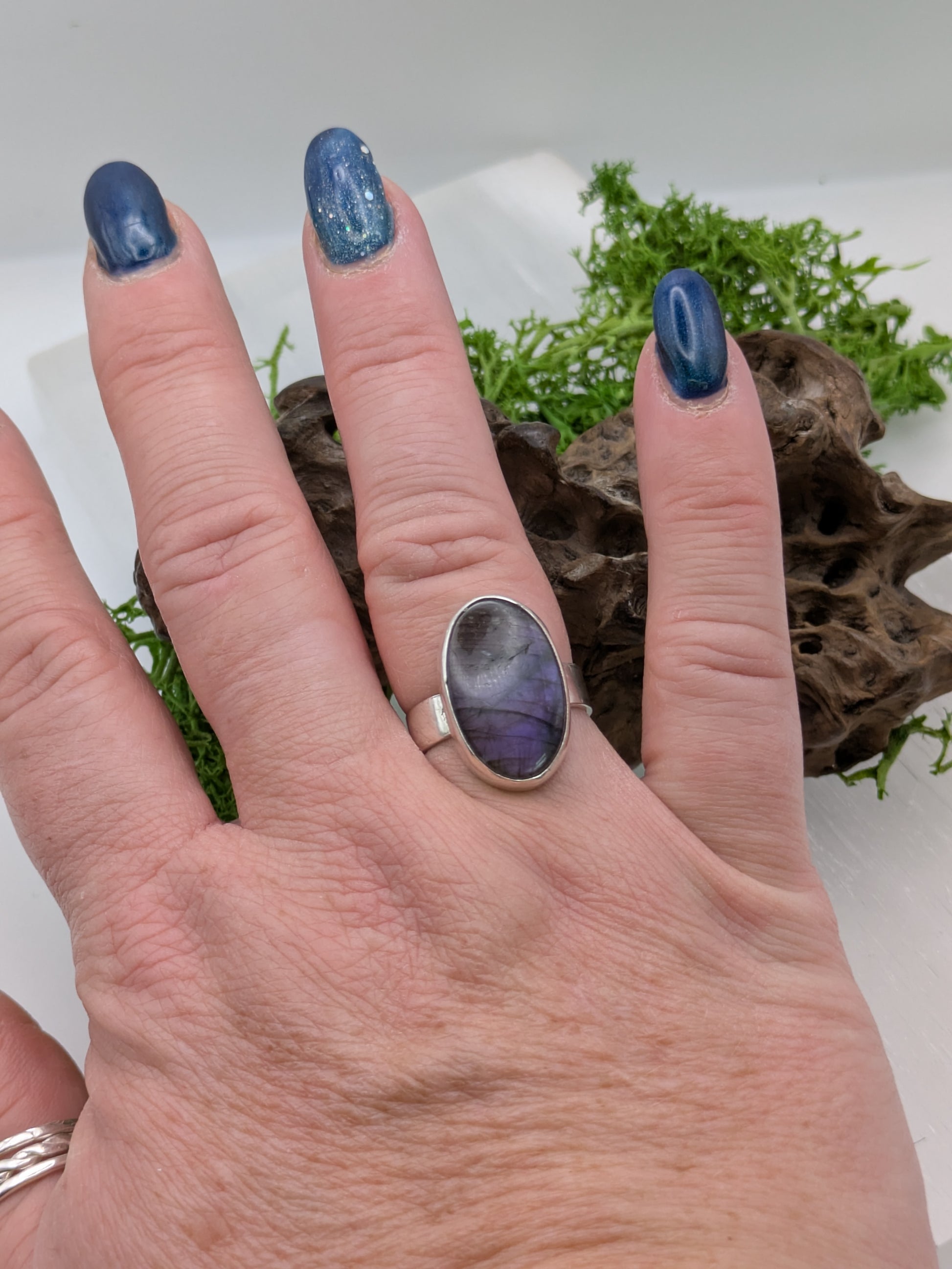 Hand wearing a silver ring with a purple Labradorite stone against a natural background