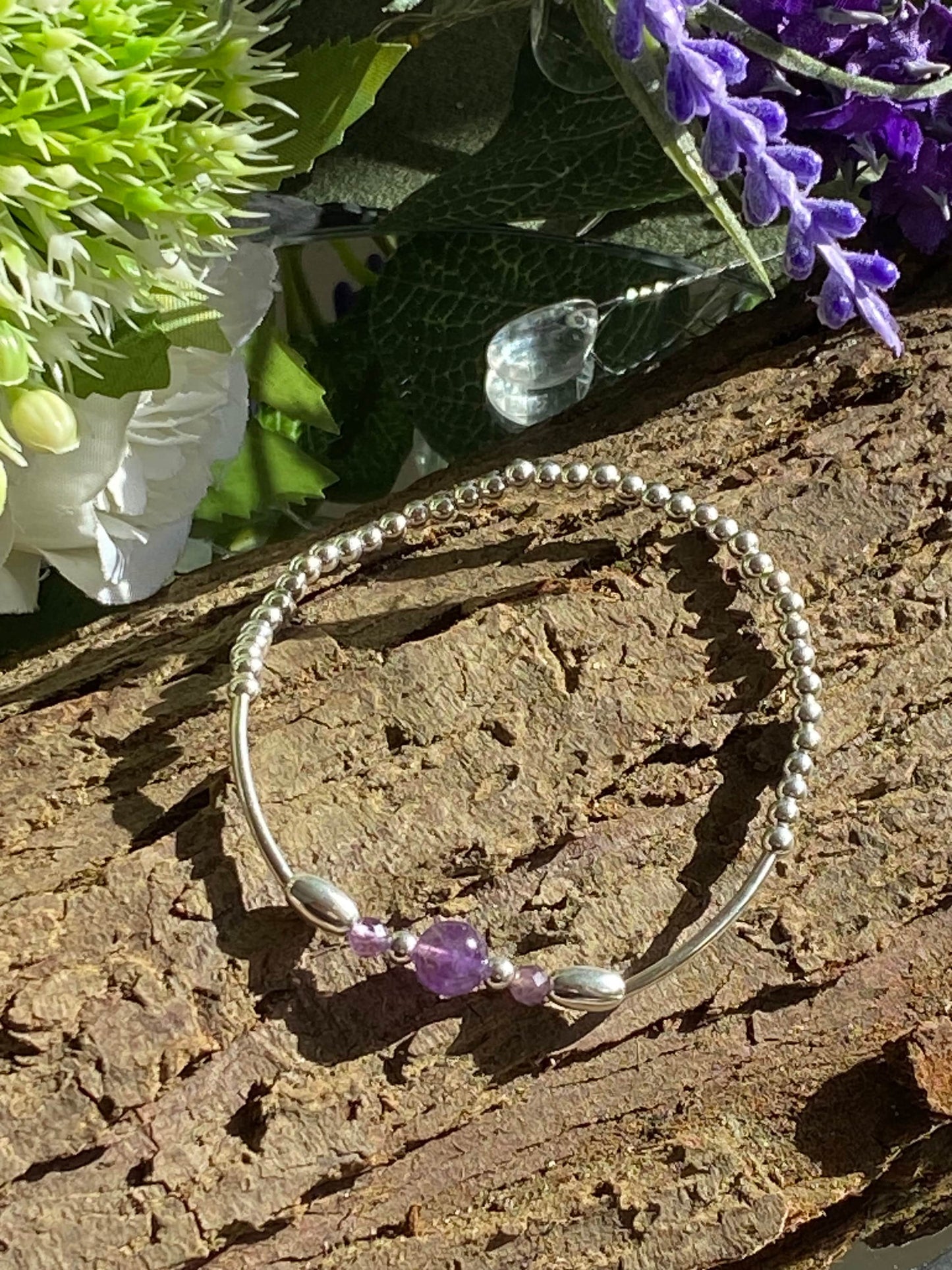 May include: Silver beaded bracelet with a small amethyst gemstone accent. The bracelet features a delicate, flexible design and is adorned with tiny, evenly spaced beads.  The amethyst is a rich purple hue, nestled between two small silver barrel-shaped beads. The bracelet is shown on a dark brown wooden background, suggesting a natural or rustic setting.
